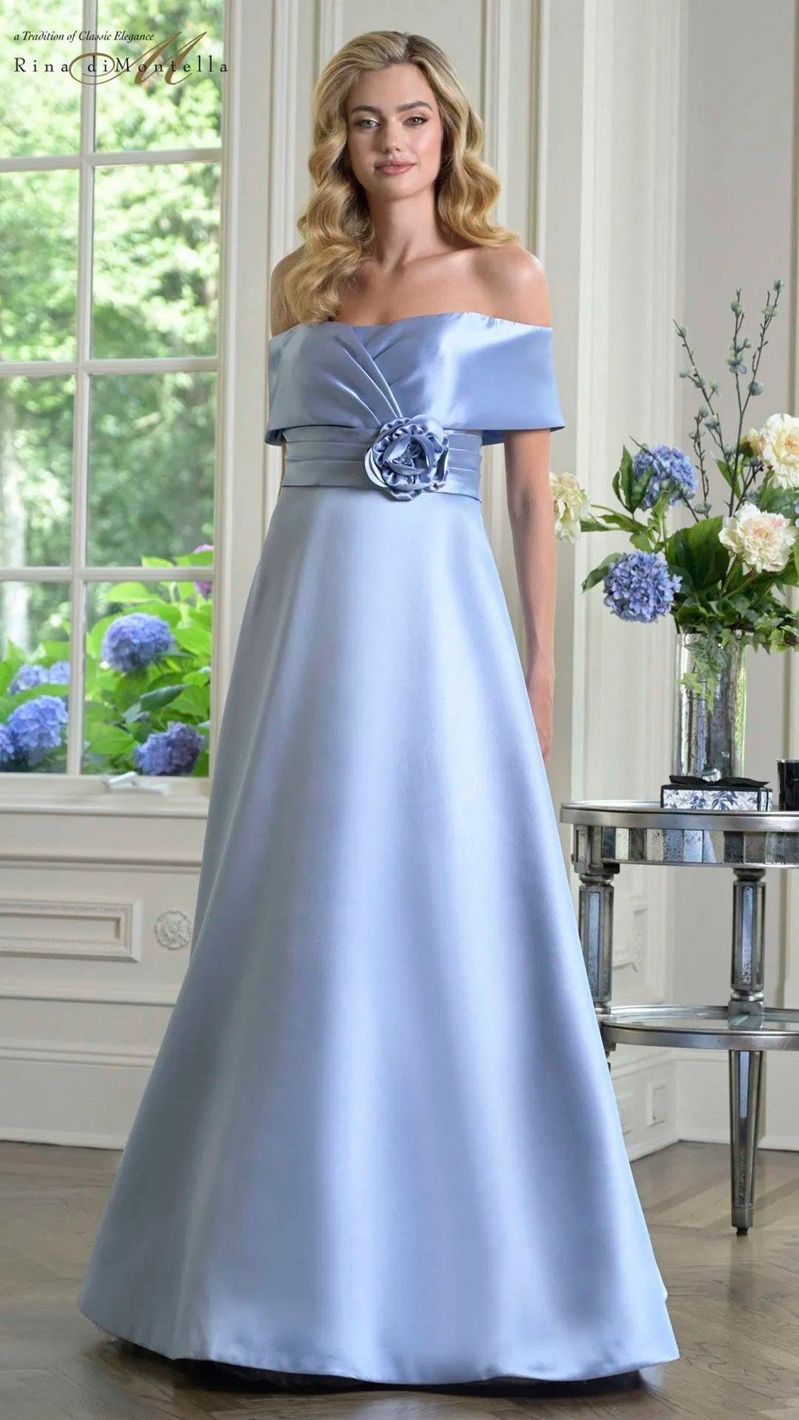 A woman stands indoors in the Rina Di Montella RD3429 long formal dress, an off-the-shoulder, floor-length sky blue gown with a floral accent at the waist; flowers and a window are visible in the background.